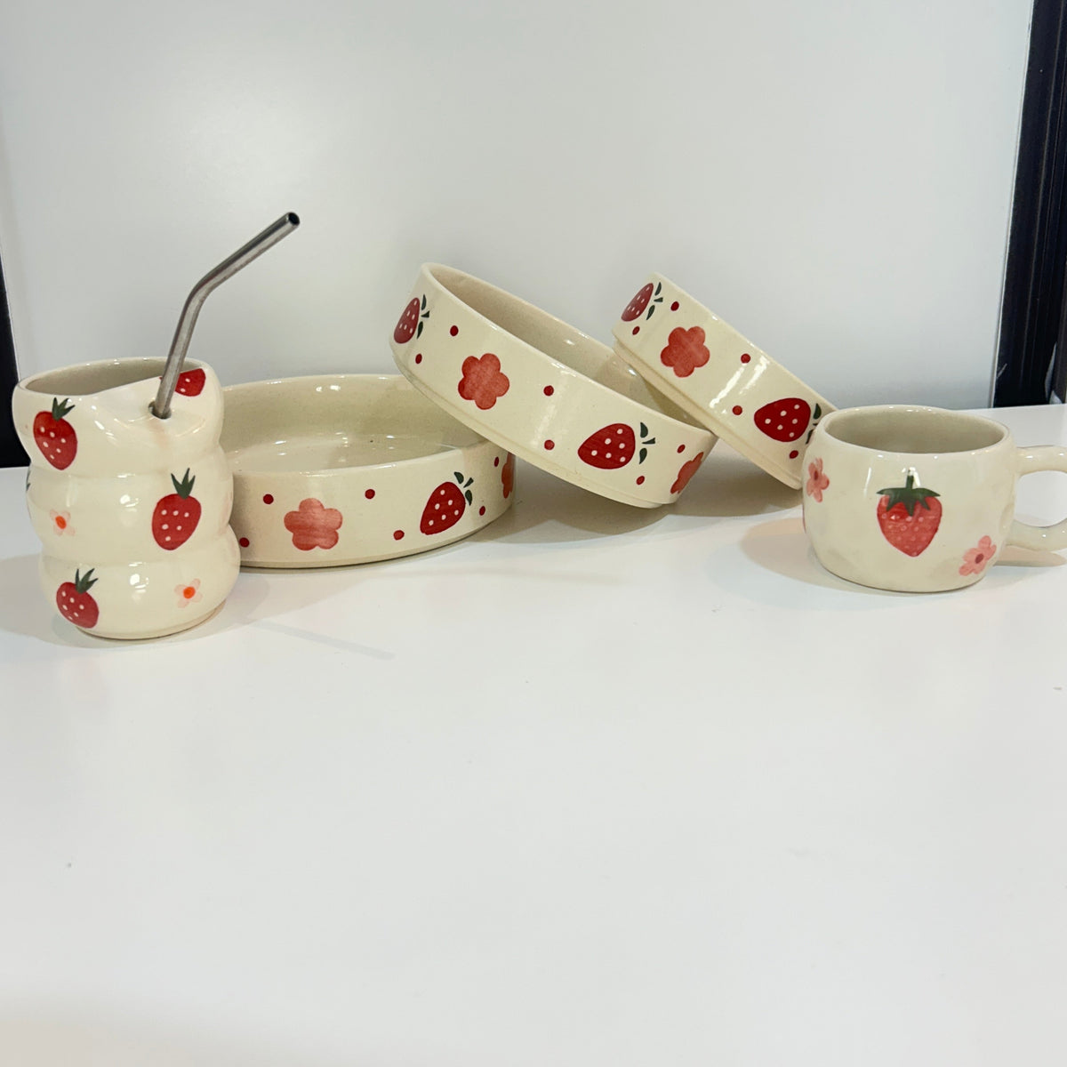 A set of ceramic tableware with a strawberry pattern, including a small handled mug, a bowl, a shallow dish, and a small cup with a metal straw, arranged on a white surface against a white background.